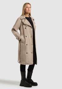 Donna che indossa un trench beige doppiopetto, un abbigliamento nero e stivali neri da combattimento con lacci, in piedi contro uno sfondo semplice.