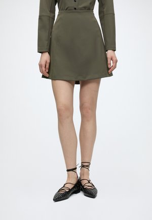 Femme portant une mini-jupe vert olive et un haut assorti à manches longues, accompagné de ballerines noires cloutées à lacets sur fond blanc.