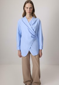 Világoskék, hosszú ujjú, ívelt szegélyű ing, laza bézs nadrággal párosítva. Simma textúra, minimalista dizájn.