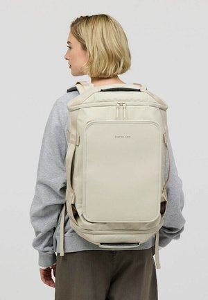 Sac à dos beige au design structuré, avec des bretelles ajustables, une poche zippée à l'avant et une texture de tissu lisse. Les accents incluent un branding minimal.