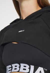Melna cropped kapuce ar neapstrādātu apakšmalu un mazu baltu NEBBIA logotipu, kas pārvalkāta virs tumša sporta krūštura ar izteiktu zīmolu joslu.