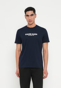Navy zilā kokvilnas T-kreklā ir apaļa kakla izgriezuma un īsām piedurknēm, ar baltu "Calvin Klein Established 1968" logotipu iegravētu priekšpusē.