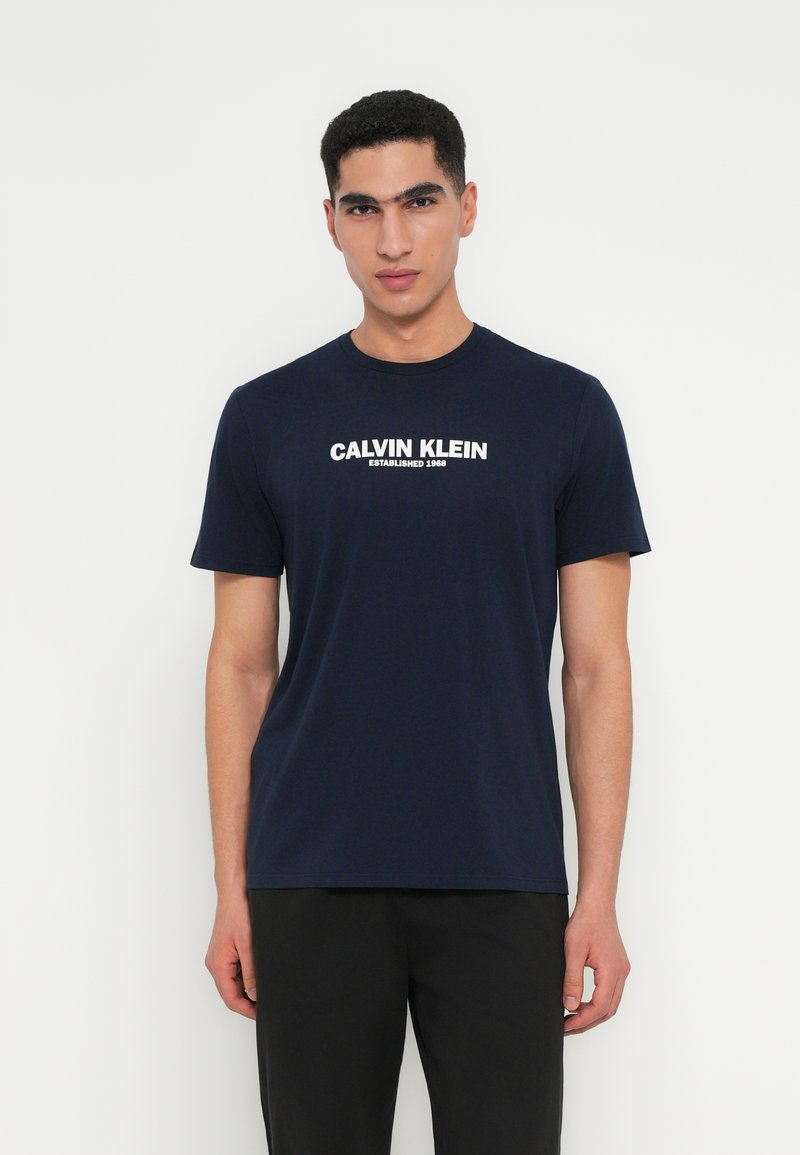 Navy zilā kokvilnas T-kreklā ir apaļa kakla izgriezuma un īsām piedurknēm, ar baltu "Calvin Klein Established 1968" logotipu iegravētu priekšpusē.