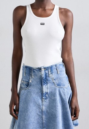 Débardeur blanc côtelé avec une petite étiquette noire, associé à une jupe en denim bleu clair dotée de poches et d'une fermeture à boutons.