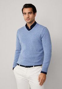 Světle modrý svetr s výstřihem do V z jemného pleteného materiálu, s žebrovanými manžetami a lemem, nošený přes tmavou košili na knoflíky a krémové kalhoty.