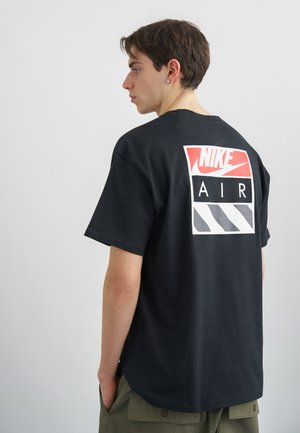 Joven vistiendo una camiseta negra Nike Air con un logo en rojo, blanco y gris en la espalda, de pie de perfil con las manos en los bolsillos de unos pantalones verdes.