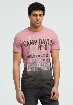 Mann mit kurzen lockigen Haaren trägt ein pink-schwarzes Farbverlauf-T-Shirt mit großem weiße und schwarzen Text und steht vor einem schlichten Hintergrund.