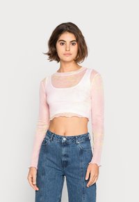 Blusa longa transparente de manga longa em rosa claro com padrão floral, corte cropped e bainha com ziguezague. Combinada com jeans de cintura alta.