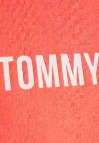 Korálová tkanina s texturovaným vzorem má velké bílé nápisy "TOMMY" umístěn�é na střed povrchu.