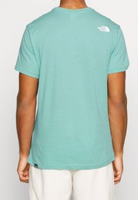 T-shirt z krótkim rękawem w kolorze teal, wykonany z miękkiego materiału. Posiada małe białe logo na górnej części pleców oraz okrągły dekolt.