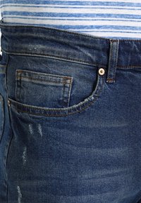 Blå denimjeans med framficka för mynt, bälteshällor och en vit-och-blå randig topp synlig ovanför midjan.