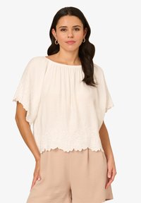 Blusa bianca ricamata con scollatura rotonda e maniche ampie; presenta motivi floreali all'orlo. Abbinata a pantaloni corti beige chiaro.