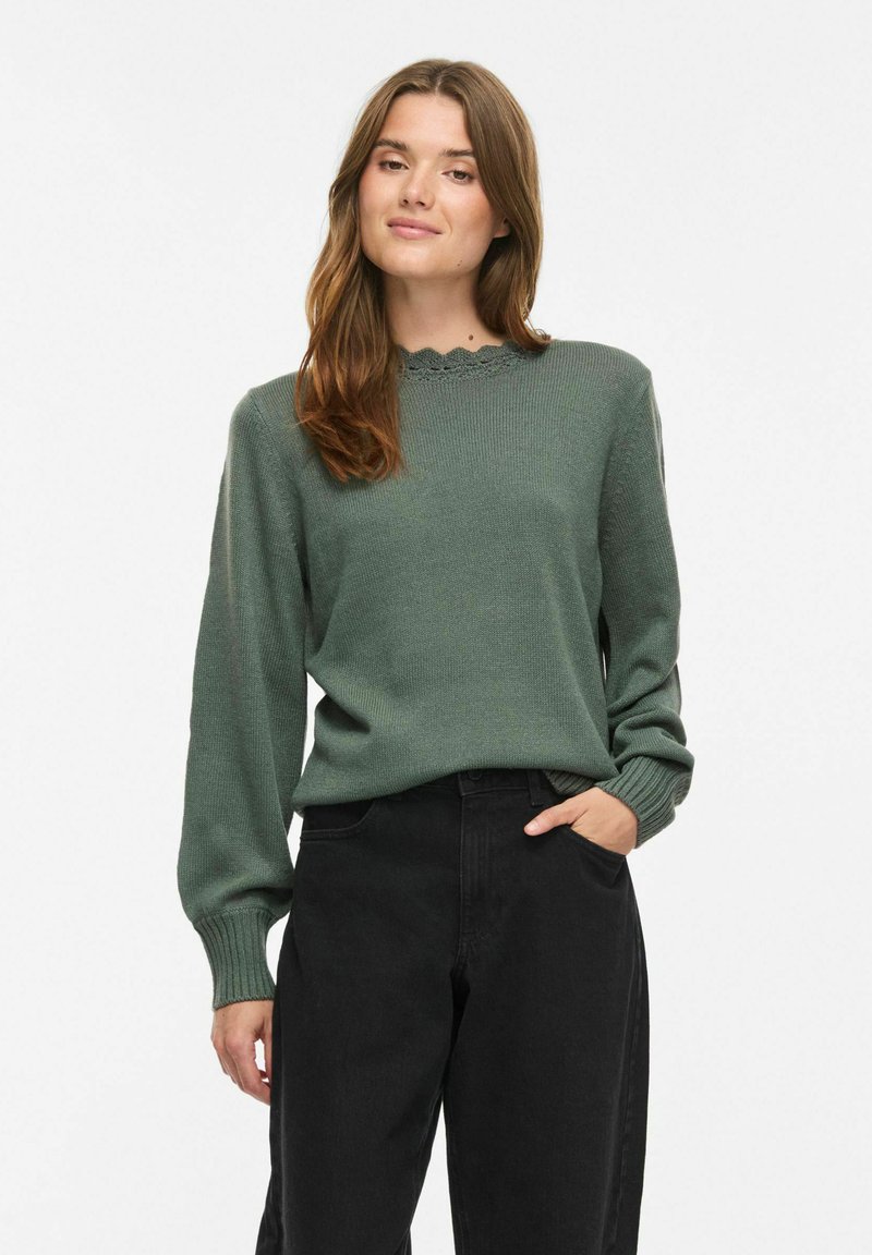 Femme aux cheveux bruns portant un pull vert à manches longues et un pantalon noir, debout, une main dans la poche, sur fond blanc.