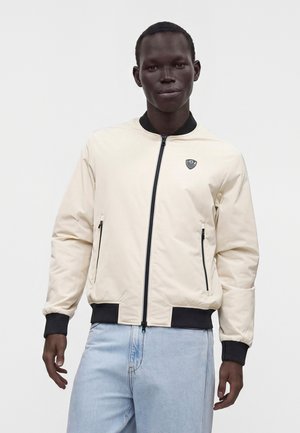 Hombre con una chaqueta bomber beige clara con cremallera, puños y cuello negros, combinado con vaqueros azul claro, de pie frente a un fondo liso.
