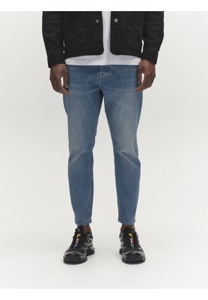 Férfi világoskék slim-fit farmert, fekete dzsekit, fehér inget és fekete sportcipőt színes részletekkel visel, fehér háttér előtt áll.