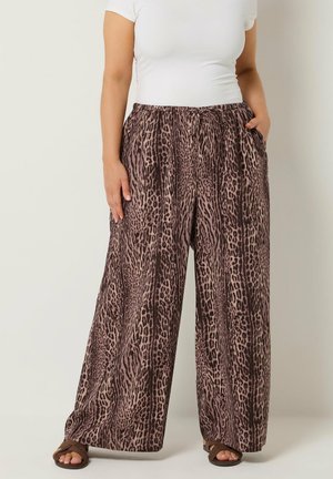 Frau trägt weit geschnittene Hose mit braunem Animal-Print, weißes eng anliegendes Kurzarmshirt und braune Sandalen mit offenem Zehenbereich.