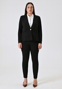 Zwarte op maat gemaakte blazer met twee voorzakken en een sluiting met één knop, gecombineerd met zwarte slim-fit broek en donkerrode hoge hakken.