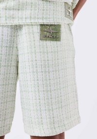 Groene en witte gebloemde shorts van textuurstof. Bevat een zichtbaar logo-patch in het groen aan de voorkant. Rechthoekige vorm met een rafelige zoom.