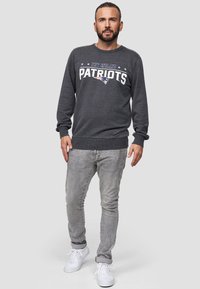 Sudadera gris con el texto y el logo de "New England Patriots", cuello redondo, puños acanalados, combinada con jeans gris claro y zapatillas blancas.