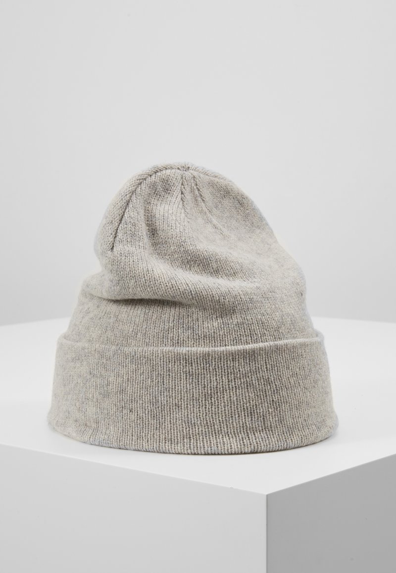 Bonnet en laine gris clair tricoté avec un bord retroussé, présenté sur un socle blanc sur un fond uni gris clair.