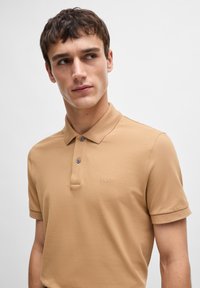 Polo de cor tan feito de tecido texturizado, apresentando uma pala de botão dupla, logótipo bordado e mangas curtas com punhos rolados.