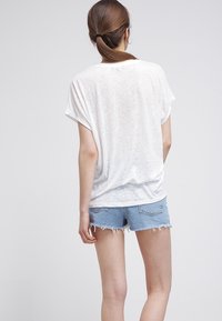 Camiseta blanca de corte holgado con mangas enrolladas, textura semitransparente, combinada con pantalones cortos de mezclilla azul claro con dobladillos deshilachados.