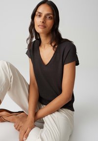 Top nero a maniche corte con scollatura a V e finitura texturizzata. Modella che indossa pantaloni beige a gamba larga e sandali piatti. Sfondo neutro.