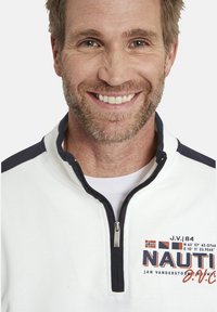 White zip-up sweatshirt featuring a navy collar and accents, with "NAUTI" logo and multi-colored text on the left side. Smooth texture.