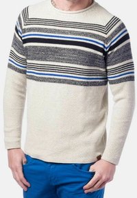 Homme portant un pull en maille crème avec des rayures horizontales noires, grises et bleues, assorti à un pantalon bleu vif, les mains dans les poches.
