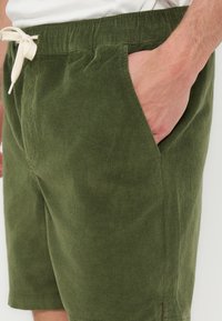 Κοντινό πλάνο ενός ατόμου που φοράει βερμούδα από κοτλέ ελιάς με λευκό κορδόνι, το χέρι ακουμπάει στην μπροστινή τσέπη.