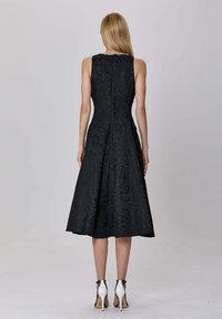 Femme aux longs cheveux blonds portant une robe midi noire texturée sans manches et des sandales blanches à talons hauts, se tenant de dos sur un fond uni.