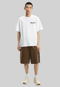 T-shirt bianca in cotone con stampa grafica nera sul petto, abbinata a pantaloni corti marroni fino al ginocchio e sneakers bianche con dettagli beige.
