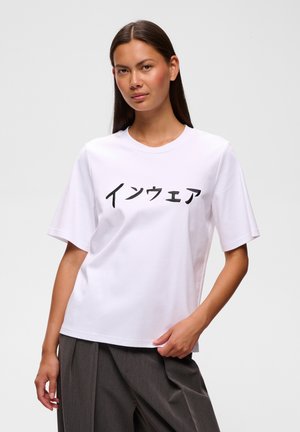 Femme portant un t-shirt blanc avec des caractères japonais, associé à un pantalon ample gris foncé, debout devant un fond clair uni.