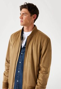 Bruine bomberjack met ribgebreide manchetten en kraag, voorzien van een ritssluiting en een zachte textuur, gelaagd over een denim overhemd.