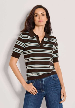 Frau mit dunklen Haaren, die ein braun-hellblau gestreiftes kurzärmeliges Poloshirt und blaue Jeans trägt, steht mit einer Hand in der Hüfte.