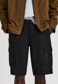 Mann in schwarzen Cargo-Shorts, einem hellgrauen Hemd, das in die Hose gesteckt ist, und einer braunen Jacke mit den Händen in den Taschen.
