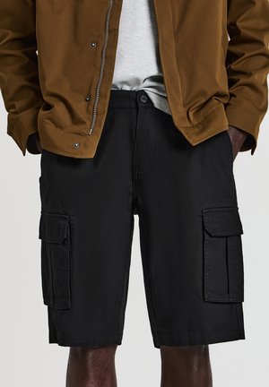 Man wearing black cargo shorts, a light grey shirt tucked in, and a brown jacket with hands in pockets.