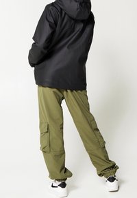 Schwarze wasserdichte Jacke mit Kapuze, kombiniert mit lockeren olivegrünen Cargo-Hosen mit Cargo-Taschen und weißen Sneakers mit schwarzen Akzenten.