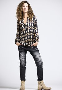 Schwarze und goldene gemusterte Bluse mit tiefem V-Ausschnitt, kombiniert mit gewaschenen, dunklen Jeans und beigefarbenen Stiefeletten mit Reißverschlüssen.