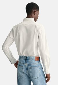 Chemise blanche à manches longues avec col à boutons, couture au dos ; associée à un jean en denim bleu clair avec un patch en cuir brun.
