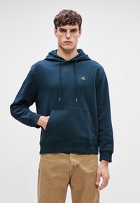 Felpa con cappuccio blu navy in morbido cotone, dotata di tasca a marsupio frontale, cappuccio regolabile con cordino e un piccolo logo dorato sul petto.