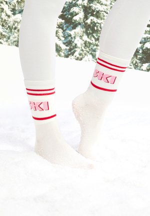 Witte skisokken tot aan de knie met rode strepen en de tekst "SKI" op de boord, geplaatst in een besneeuwd terrein, met de nadruk op de zachte textuur en het sportieve ontwerp.