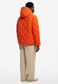 Giubbotto arancione imbottito con cappuccio, caratterizzato da motivi di cucitura diagonale. Indossato con pantaloni beige e scarpe di colore chiaro.