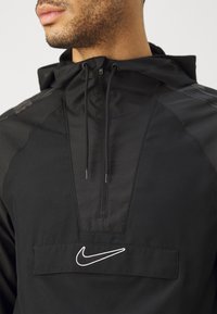 Zwarte hoodie met een hoge kraag, voorzien van een kwartritssluiting en een wit Nike-logo op de voorkant. Gelaagd materiaal aan de zijkanten.