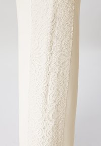 Panel de tela color marfil con un intrincado patrón de encaje floral y bordes con ondas sobre un fondo liso.