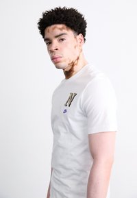 Witte T-shirt met korte mouwen, voorzien van een gestructureerde, betitelde letter "N" in zwart en geel, met een klein paars Nike-logo op de borst.