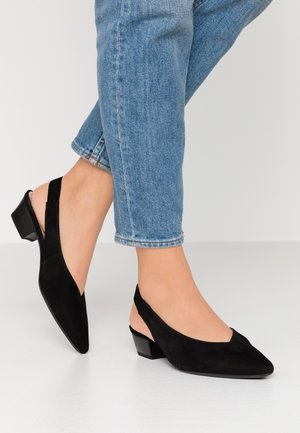 Kobieta nosząca czarne sandały typu slingback na niskim obcasie ze spiczastymi noskami oraz krótkie niebieskie jeansy na białej podłodze.