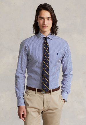 Mężczyzna o długich ciemnych włosach, ubrany w niebiesko-białą pasiastą koszulę, granatowy krawat w wzory, beżowe spodnie i brązowy pasek, stojący na jednolitym tle.