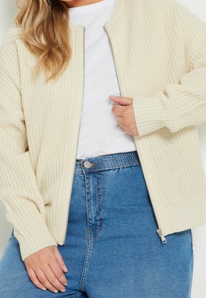 Vrouw in een crèmekleurige gebreide ritsjack over een wit shirt en hoge blauwe spijkerbroek, met een hand die de rits aanpast.