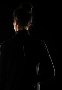 Sort atletisk jakke med høj krave, der har en reflekterende vertikal detalje på ryggen. Glat tekstur og tætsiddende design.